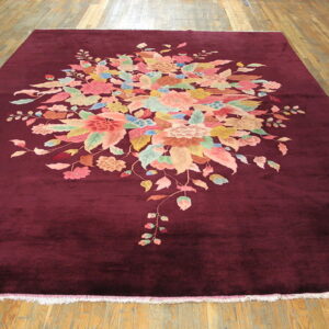 Plush maroon rug with a dense central pattern of pink, green, and blue flowers on a wood floor.