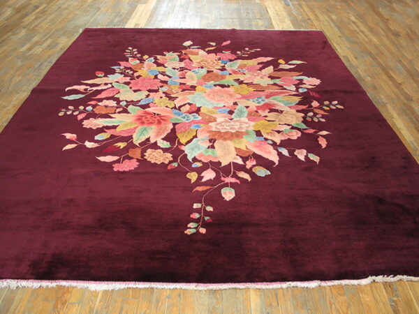 Plush maroon rug with a dense central pattern of pink, green, and blue flowers on a wood floor.