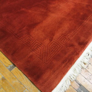 Reddish-orange low-pile rug showing a carved geometric border and cream fringe on distressed wood flooring.