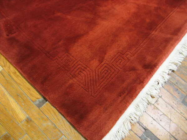 Reddish-orange low-pile rug showing a carved geometric border and cream fringe on distressed wood flooring.