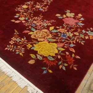 Deep red rug with colorful floral vine pattern and white fringe on light wood flooring.