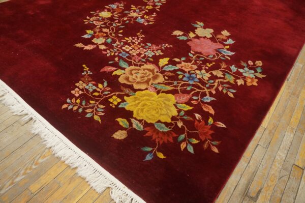 Deep red rug with colorful floral vine pattern and white fringe on light wood flooring.
