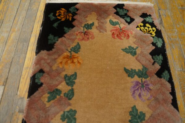 Floral patterned rug with a tan center and stepped geometric border on worn yellow wood flooring.