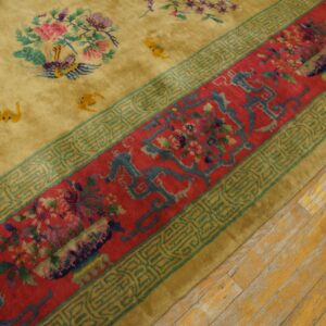A high-pile gold rug with floral details and a wide red border rests on worn yellow wood flooring.