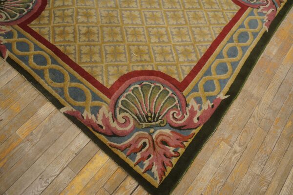 Low-pile rug corner showing gold lattice field and blue border with pink shell scrollwork on wood floors.
