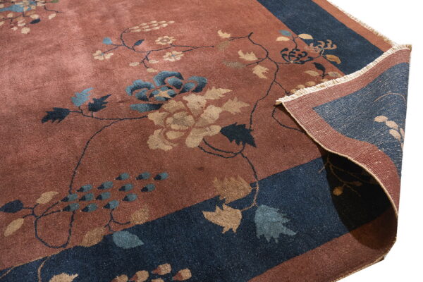 A rusty-red rug with blue and gold floral vines has its deep navy border corner folded up.