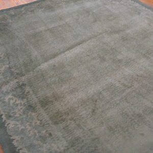 Muted sage rug with a decorative border sits on planks of warm orange hardwood flooring.