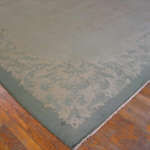 Low-pile sage green rug with a tonal floral border rests on worn hardwood floors.