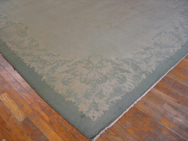 Low-pile sage green rug with a tonal floral border rests on worn hardwood floors.