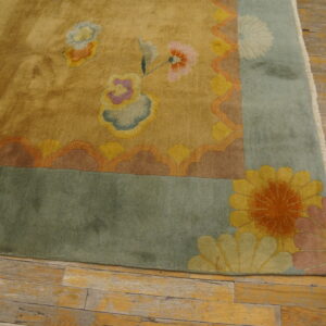 Low pile yellow rug corner with a blue-green border and multicolored floral designs on worn wood flooring.