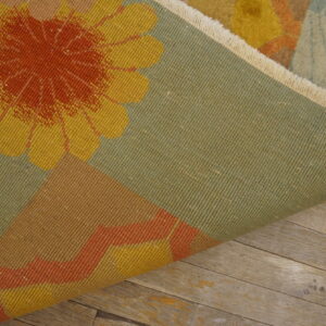 Yellow and orange floral pattern rug folded back, showing the tight green backing on weathered wood flooring.