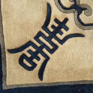 Close-up of a rug featuring a tan field with dark blue and gray geometric and scrolling stylized patterns.