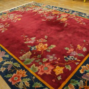 A deep red rug featuring a contrasting navy blue floral border is displayed on light hardwood floors.