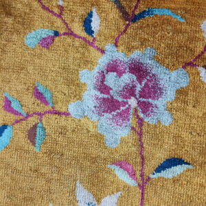 Textured golden rug with a pink border and a large floral design in blue, magenta, and brown tones.
