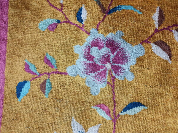 Textured golden rug with a pink border and a large floral design in blue, magenta, and brown tones.