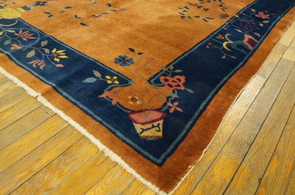 Burnt orange rug with a navy blue border displaying colorful floral and vase motifs on wood flooring.