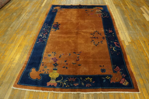 Plush orange rug with a navy blue border featuring multicolored floral and traditional decorative patterns on wood planks.