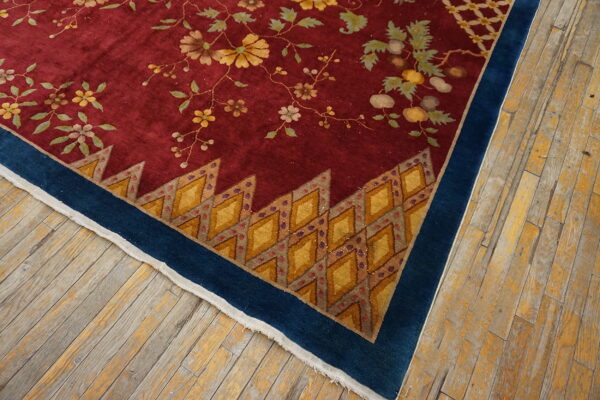 Maroon rug corner showing yellow vines, a jagged diamond inner border, and a solid blue outer border on worn wood floors.