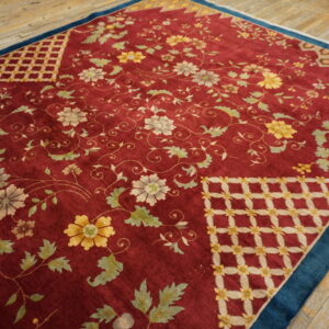 Low-pile red rug with dense floral patterns, geometric corners, and a dark blue border on wood planks.