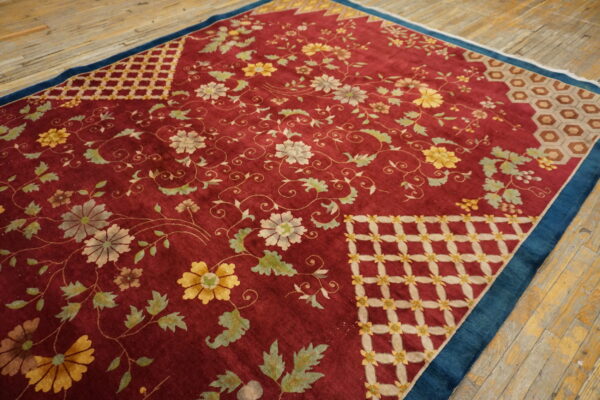 Low-pile red rug with dense floral patterns, geometric corners, and a dark blue border on wood planks.