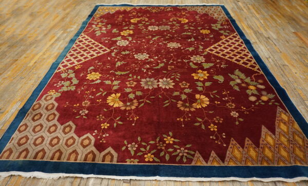 Red floral rug with blue borders and geometric panels rests on light worn wooden floorboards.