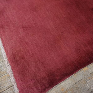 A deep red solid rug with visible pile variation rests on rustic gray wooden floorboards.