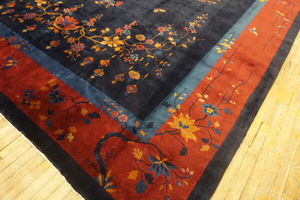 Ornate dark blue rug featuring multicolor floral branches and a wide rust red border on wood flooring.