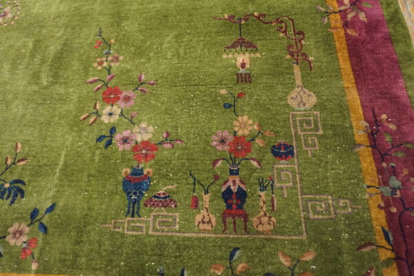 Detailed view of a moss green rug with stylized floral motifs, decorative objects, and a maroon and gold border.