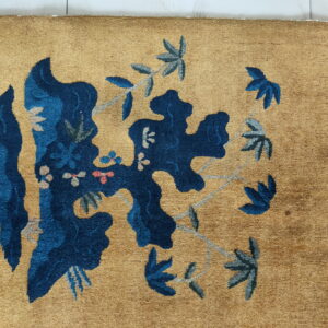 A golden tan rug featuring a large, stylized dark blue and gray floral vine pattern.