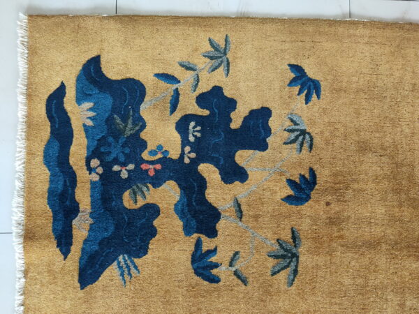 A golden tan rug featuring a large, stylized dark blue and gray floral vine pattern.