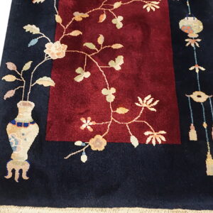 Navy rug with burgundy center panel, featuring cream vase, floral vines, and hanging tassel motifs.