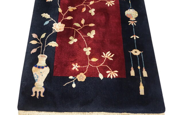 Navy rug with burgundy center panel, featuring cream vase, floral vines, and hanging tassel motifs.