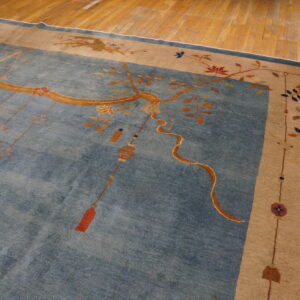 Blue rug with tan border features gold branches, hanging tassels, and floral motifs on wood flooring.