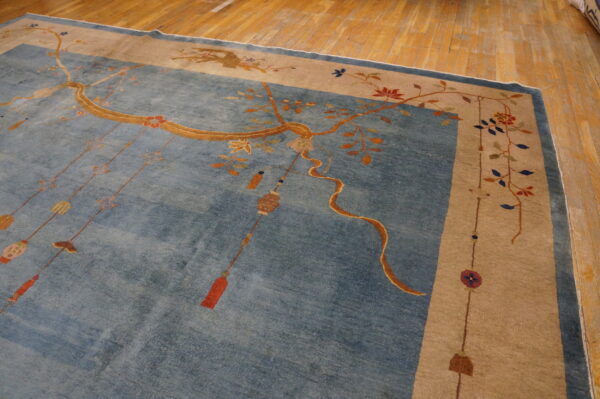 Blue rug with tan border features gold branches, hanging tassels, and floral motifs on wood flooring.