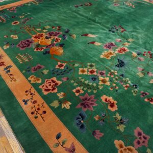High-pile green rug with multicolor floral motifs and an orange border rests on a worn wood floor.