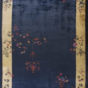Dark blue rug featuring a sheen and a wide beige border decorated with intricate flowering branches and vases.