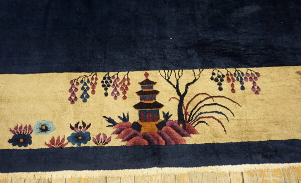 High-pile navy rug border with a beige scenic pattern of a pagoda, flowers, and foliage resting on wood.