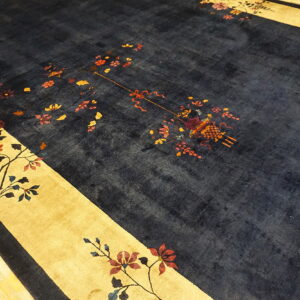 A plush navy blue rug with gold floral borders and scattered multicolored motifs rests on light hardwood flooring.