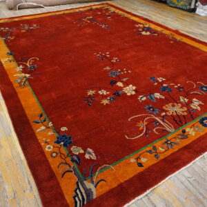Ornate red rug with a wide orange border and colorful floral motifs displayed on distressed wood planks.