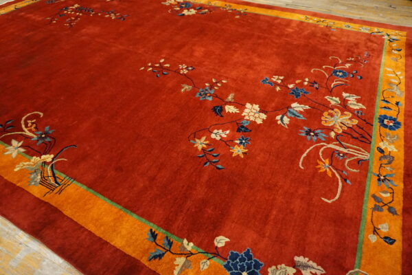 A deep red, high-pile rug displays delicate blue and cream floral vines surrounded by a bright orange border.