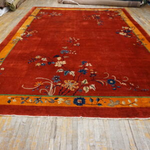 Red area rug featuring a wide orange border and delicate floral designs in blue and tan displayed on worn plank flooring.