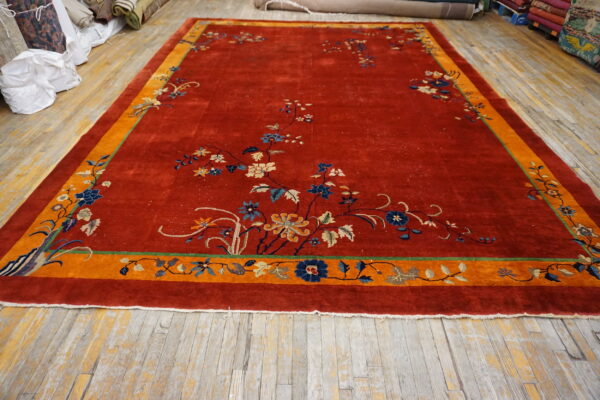 Red area rug featuring a wide orange border and delicate floral designs in blue and tan displayed on worn plank flooring.