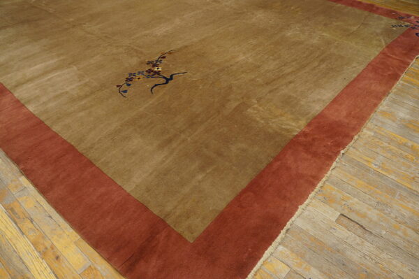Tan low-pile rug with a single floral motif and thick rust-colored border on worn wooden floor.
