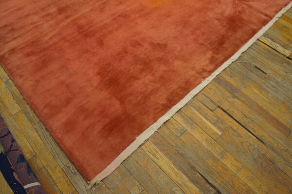 A solid rust-orange rug with high sheen texture rests on distressed, diagonal hardwood flooring.