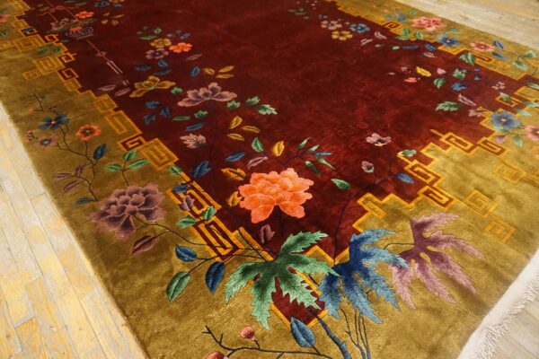 A plush burgundy and gold rug with an all-over floral pattern and geometric key border on wood flooring.