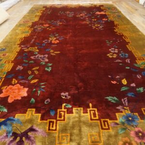 A deep red rug featuring multicolored floral designs and an olive border with a continuous geometric fret pattern.