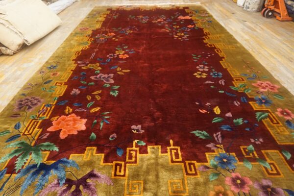 A deep red rug featuring multicolored floral designs and an olive border with a continuous geometric fret pattern.