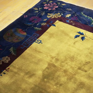 Gold rug corner featuring a navy blue border detailed with complex floral and geometric motifs on wooden flooring.