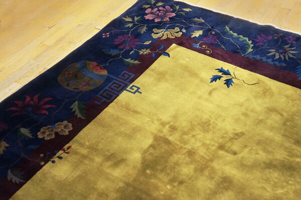 Gold rug corner featuring a navy blue border detailed with complex floral and geometric motifs on wooden flooring.