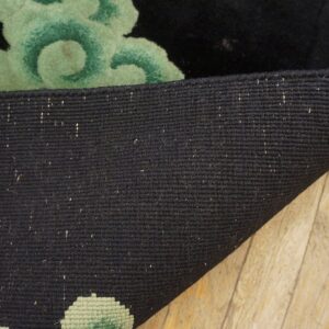 Black woven rug backing is curled to show the green floral pattern and black pile on wood flooring.